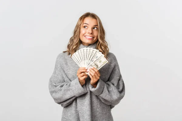 Portret van dromerig schattig meisje in grijze trui, kijken naar de linker bovenhoek bedachtzaam en glimlachend, met geld, witte achtergrond — Stockfoto