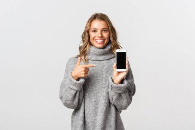 Gri kazaklı çekici sarışın kızın portresi cep telefonu ekranını işaret ediyor ve gülümsüyor.