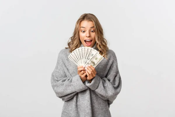 Изображение возбужденной блондинки в сером свитере, счастливо смотрящей на деньги в руках, стоящей на белом фоне — стоковое фото