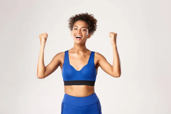 Concept of sport and workout. Happy african-american fitness girl cheering, fist pump like a champion, celebrating victory, triumphing against white background