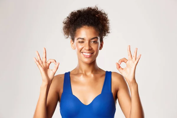 Close-up of athletic african-american fitness woman in blue workout clothing, showing okay signs and winking, smiling satisfied, white background — Stock Photo, Image