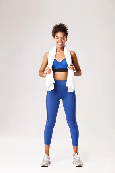 Comprimento total de mulher fitness saudável sorridente em uniforme azul, segurando toalha e olhando satisfeito após bom treino, em pé sobre fundo branco — Fotografia de Stock