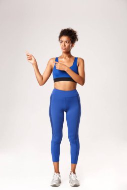 Çekici Afro-Amerikan sporcu kadın üzgün görünüyor, parmaklarını sola gösteriyor ve somurtuyor, hayal kırıklığını ya da hoşnutsuzluğunu ifade ediyor, mavi spor giyim, beyaz arka plan