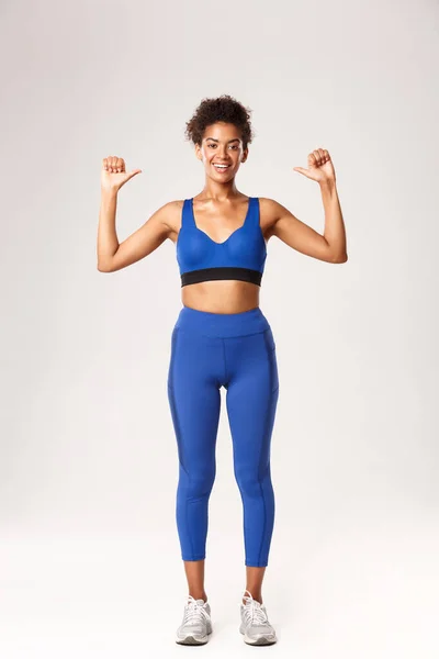 Longitud completa de la deportista afro-americana confiada feliz en traje azul del deporte, señalándose con sonrisa alegre orgullosa, mostrando progreso del entrenamiento, de pie sobre fondo blanco — Foto de Stock