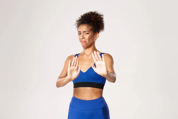 Motvillig och missnöjd afrikansk-amerikansk fitness flicka i blå sport kostym, visar stopp tecken, vägra något dåligt och grimacing från motvilja, vit bakgrund — Stockfoto