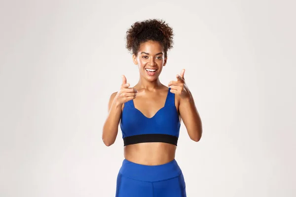 Studio shot van gelukkige Afrikaans-Amerikaanse fitness meisje in blauw uniform, wijzende vingers naar de camera en glimlachen, uitnodigen voor training, staan tegen een witte achtergrond — Stockfoto