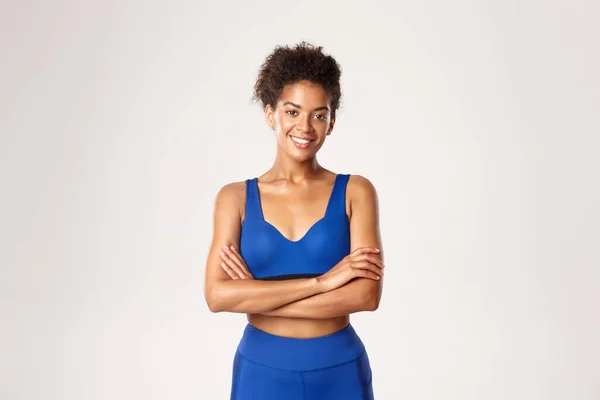 Aantrekkelijk fit Afrikaans-Amerikaans fitnessmeisje in blauwe sportkleding, armen over de borst en glimlachend, klaar voor training en oefeningen, staande tegen witte achtergrond — Stockfoto