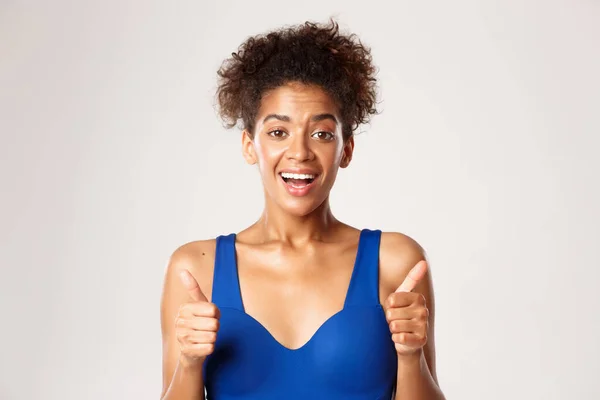 Close-up van opgewonden Afrikaans-Amerikaanse fitnessvrouw in blauwe sportbeha, met duimen-up in goedkeuring, als iets goeds, staande tegen een witte achtergrond — Stockfoto