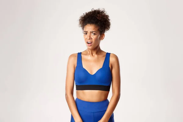 Close-up van geschokt en ontevreden Afrikaans-Amerikaanse fitness meisje, kijkt gefrustreerd naar iets slechts, staan tegen een witte achtergrond — Stockfoto