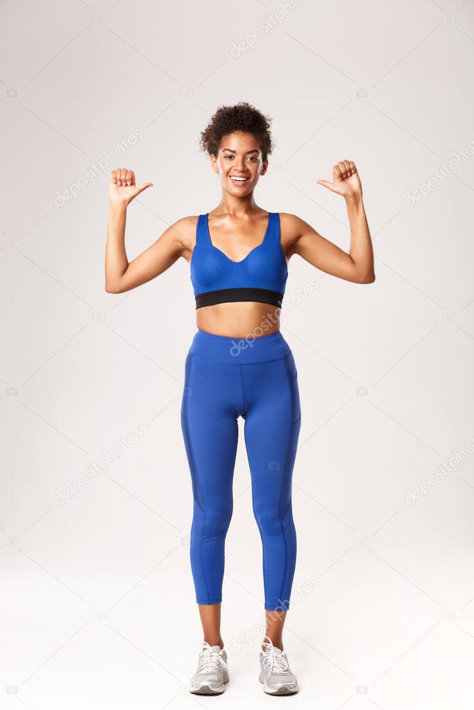 Full length of happy confident african-american sportswoman in blue sport outfit, pointing at herself with proud cheerful smile, showing workout progress, standing over white background