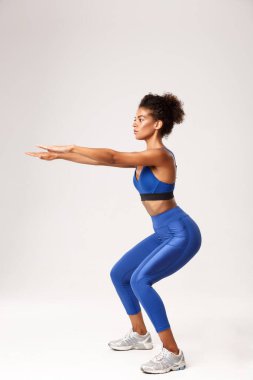 Kararlı Afro-Amerikan fitness kadınının tam boy profil çekimi, mavi spor kıyafet giymiş, elleri öne doğru uzanmış, beyaz arka plan üzerinde egzersiz