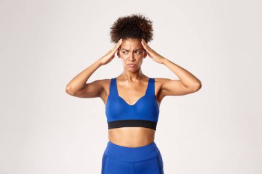 Üzüntülü Afro-Amerikan bayan sporcu, mavi spor giymiş, el ele tutuşmuş ve üzgün, baş ağrısı ile gergin, beyaz arka plan