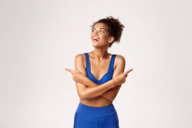 Çekici Afro-Amerikan spor giysili kadın, parmaklarını yana doğru gösteriyor ve sol üst köşeye bakıyor, mutlu görünüyor, beyaz arka plana karşı duruyor.