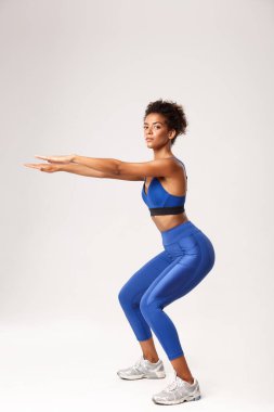 Mavi spor kostüm giymiş, çömelme egzersizleri için ellerini uzatan, beyaz arka plan üzerinden kameraya bakan güzel ve sağlıklı Afro-Amerikan kadın.