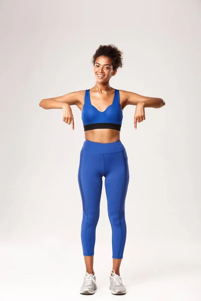 Comprimento total de treinador de fitness afro-americano feliz em sportswear azul, apontando os dedos para baixo e sorrindo, recomendo equipamento de treino, mostrando logotipo ou promo esportes, fundo branco — Fotografia de Stock