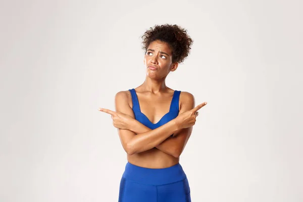 Concept van training en fitness. Trieste besluiteloze sportvrouw in blauwe sport uniform, wijzende vingers zijwaarts, maar links kijkend met teleurgesteld gezicht, witte achtergrond — Stockfoto