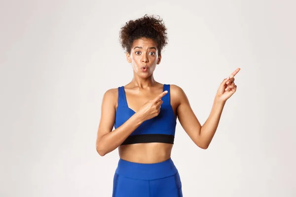 Концепція тренування та фізичної підготовки. Здивована і цікава афро-американська жінка-спортсменка в блакитному спортивному одязі, вказуючи пальці у верхньому правому куті, білий фон — стокове фото