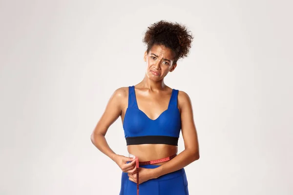 Teleurgesteld en verdrietig Afrikaans-Amerikaanse fitness meisje klagen, taille meten met tape en grimmig boos met haar lichaam, witte achtergrond — Stockfoto