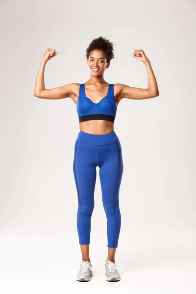 Longitud completa de fuerte y confiada chica afroamericana mostrando sus músculos, bíceps flexibles y sonriendo, demostrar el gimnasio y el progreso del entrenamiento, de pie voer fondo blanco — Foto de Stock
