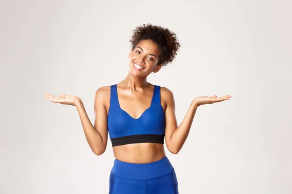 Taille-up shot van glimlachende Afrikaans-Amerikaanse fitness meisje in blauwe sport oufit, handen omhoog opzij, iets vasthouden of demonstreren, staan tegen een witte achtergrond — Stockfoto