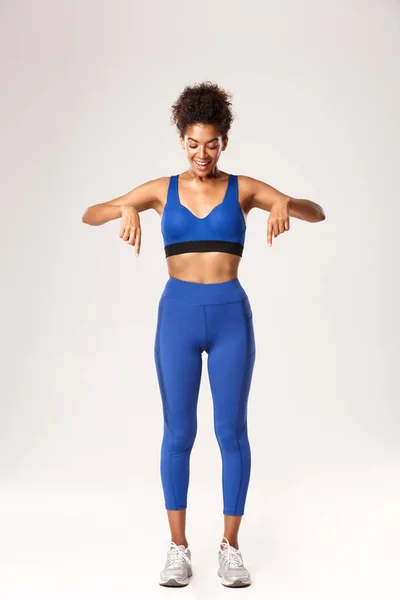 Comprimento total do atleta feminino afro-americano espantado, vestindo roupas esportivas azuis, apontando os dedos para baixo e olhando com sorriso animado, em pé sobre o fundo branco — Fotografia de Stock