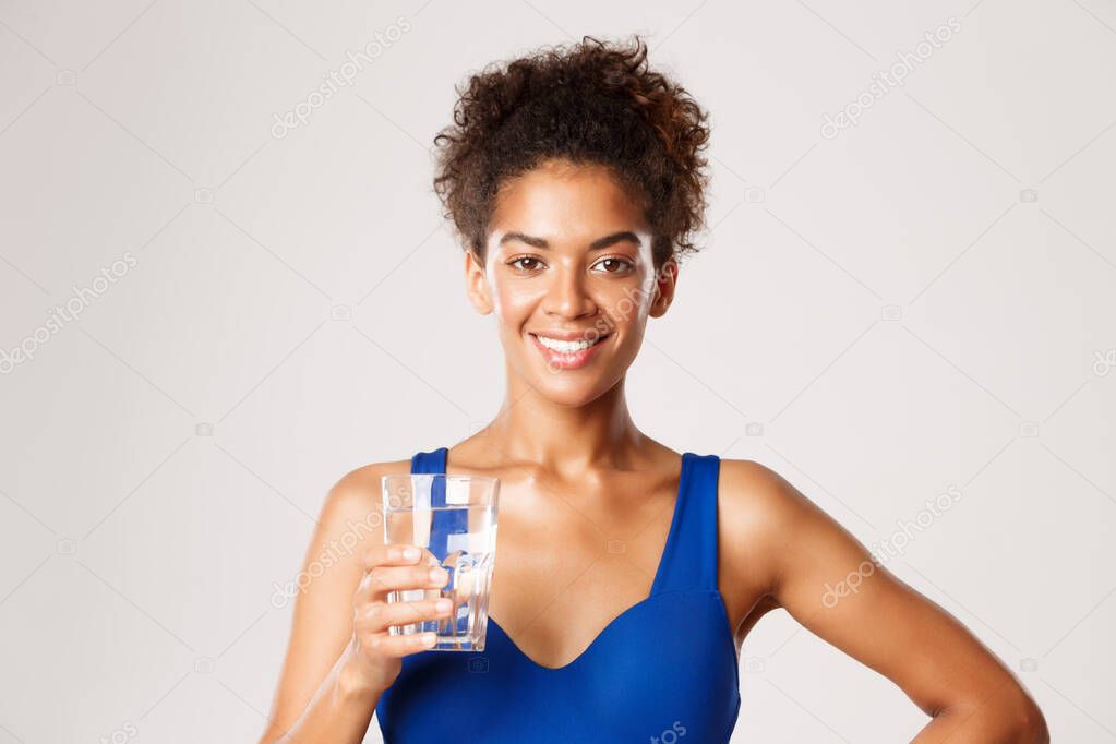 Close-up of young happy african-american fitness woman, holding glass of water and smiling pleased, standing against white background