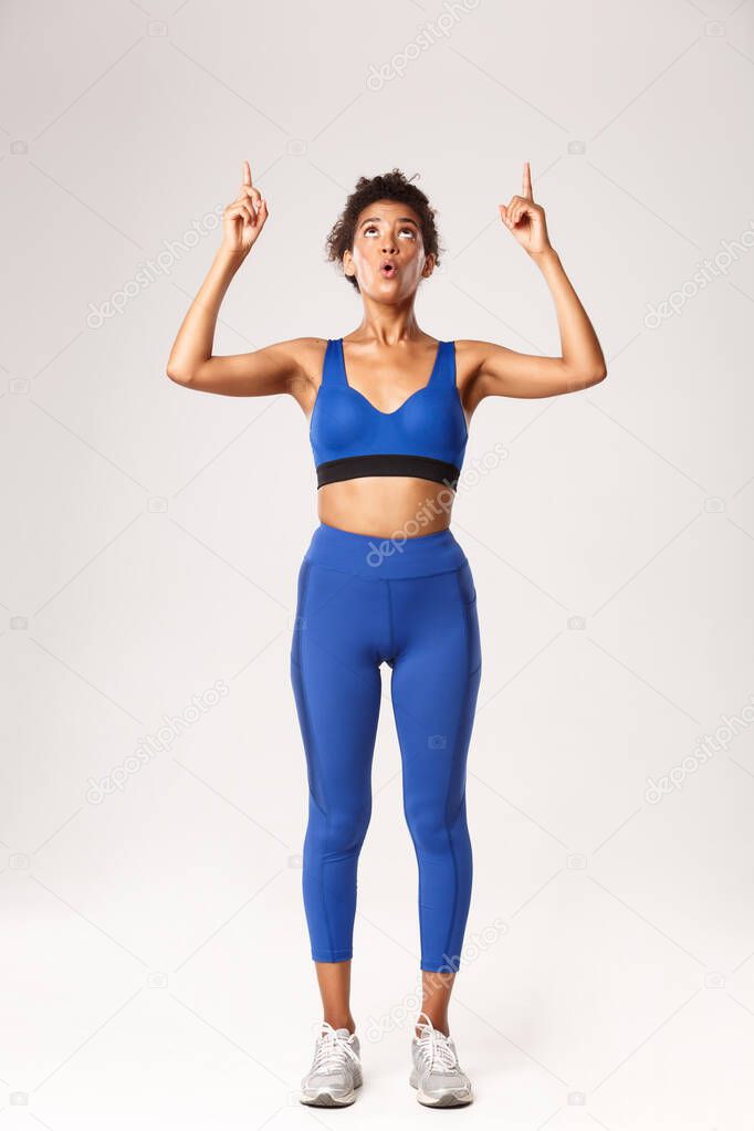 Full length of amazed african-american sportswoman, looking and pointing fingers up with wondered face, standing in blue sports costume over white background