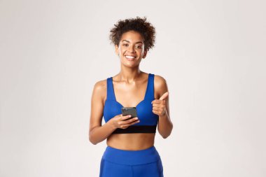 Spor ve teknoloji konsepti. Formda ve sağlıklı Afro-Amerikan bayan sporcu, cep telefonu kullanırken onayını veriyor, beyaz arka plan