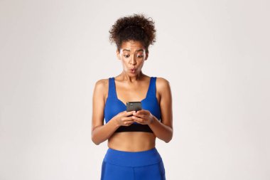 Spor ve teknoloji konsepti. Beyaz arka planda duran, akıllı telefon ekranına heyecanlı görünen, spor kıyafetli Afro-Amerikan formalı kız.