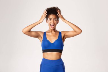Spor ve spor anlayışı. Telaşlı ve sıkıntılı Afro-Amerikan bayan sporcu çığlık atıyor, başının yanında el sıkışıyor, beyaz arka planda panikliyor.