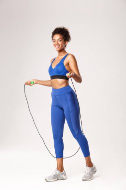 Çekici, formalı Afro-Amerikan sporcu kadın, mavi üniforma giyiyor, ip atlıyor ve kameraya gülümsüyor, beyaz arka plana karşı çalışıyor.