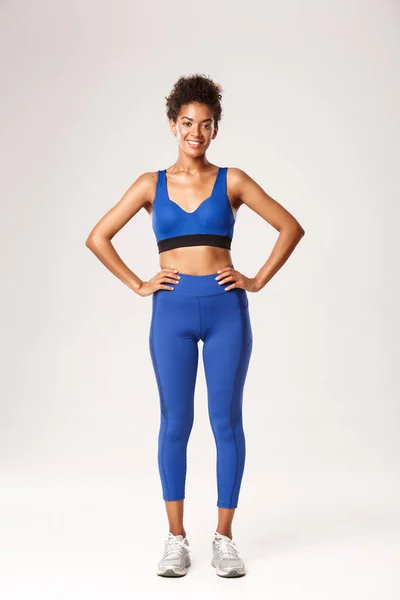 Spor giyimli, kendinden emin Afro-Amerikan bir kız, gülümseyen, kararlı bir ifadeyle el ele tutuşan, beyaz arka planda egzersiz yapan. — Stok fotoğraf