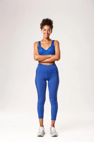Comprimento total de sorridente menina afro-americana, treinador de fitness em sportswear, sorrindo com os braços cruzados no peito, de pé pronto para o treino sobre fundo branco — Fotografia de Stock