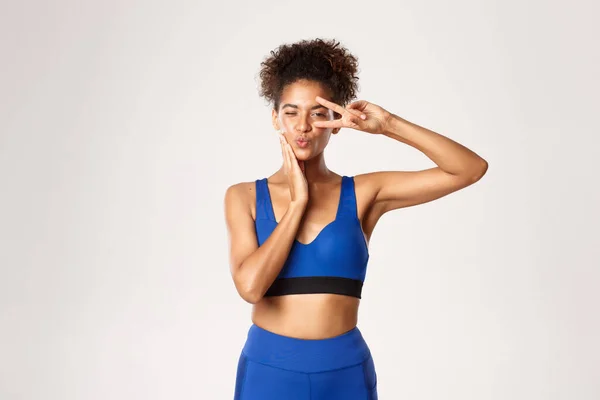 Concept van sport en training. Heerlijk Afrikaans-Amerikaans fitness meisje in blauwe sportschool kleding, tonen vrede teken en pruilen dwaas, staande over witte achtergrond — Stockfoto