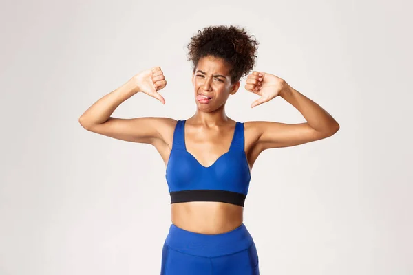 Studio skott av besviken afrikansk-amerikansk kvinnlig idrottsman, bär blå sport outfit, klagar som visar tummen ner och klibbar tunga från ogillar, vit bakgrund — Stockfoto