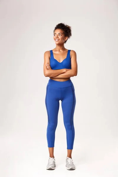 Comprimento total de forte atraente esportista afro-americano em sutiã esportivo e leggings, olhando para a esquerda e sorrindo para algo, de pé sobre fundo branco, pronto para treino — Fotografia de Stock