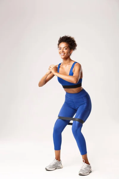 Comprimento total de mulher sorrindo apto desfrutando de fitness, vestindo roupas de esporte azul, fazendo agachamentos com banda de resistência, de pé contra fundo branco — Fotografia de Stock