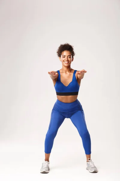 Pleine longueur de belle femme afro-américaine en tenue de sport, les mains tendues vers l'avant et faire des exercices de squats, souriant à la caméra, debout sur fond blanc — Photo