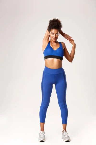 Pełna długość uśmiechu atrakcyjna afrykańsko-amerykańska fitness dziewczyna w niebieskim stroju sportowym, wyciągające ręce, rozgrzewka przed treningiem, stojąca na białym tle — Zdjęcie stockowe