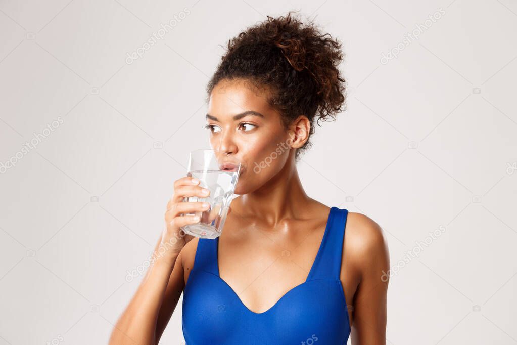 Close-up of young healthy fitness woman drinking glass of water, standing over white background in workout clothing