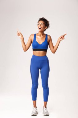 Heyecanlı ve mutlu Afro-Amerikan spor kadın, mavi spor giyer, parmaklarını yana doğru gösterir ama mutlu bir gülümseme ile sola bakar, beyaz arka plan