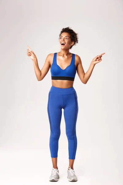 Comprimento total de animado e feliz esportista afro-americano, vestindo sportswear azul, apontando os dedos para o lado, mas olhando para a esquerda com sorriso satisfeito, fundo branco — Fotografia de Stock