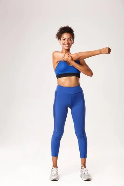 Comprimento total de bela mulher esportista afro-americana sorridente, vestindo roupas esportivas azuis, aquecimento antes do treino, mostrando exercícios de alongamento, fundo branco — Fotografia de Stock