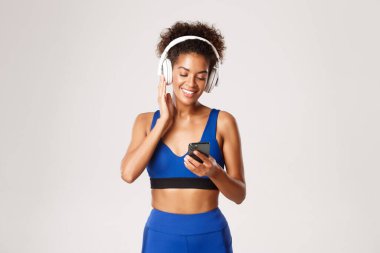 Mavi spor kıyafetli güzel bir kadın, kablosuz kulaklıkla müzik eşliğinde çalışıyor, akıllı telefon uygulaması kullanıyor, beyaz arka plana karşı duruyor.