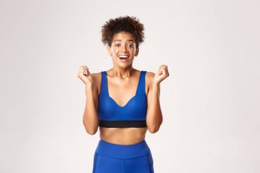 Spor ve spor anlayışı. Heyecanlı ve müteşekkir Afro-Amerikan fitness kadını kazanıyor, rahatlamış görünüyor, yumruklarını sallıyor, zevk alıyor, amacına ulaşıyor, beyaz arka planda duruyor.