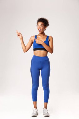 Mavi spor kostümlü afro-amerikan bayan sporcu, parmaklarını sola uzatıp şaşkınlıkla bakıyor, spor ve spor hakkında bir şeyler gösteriyor, beyaz arka plan