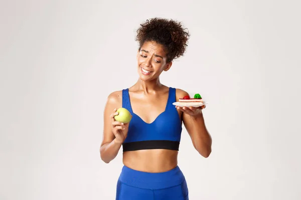 Здоровый образ жизни и спортивная концепция. Нерешительная и мрачная африканско-американская фитнес-женщина на диете, с соблазном смотреть на торт, но есть яблоко, белый фон — стоковое фото