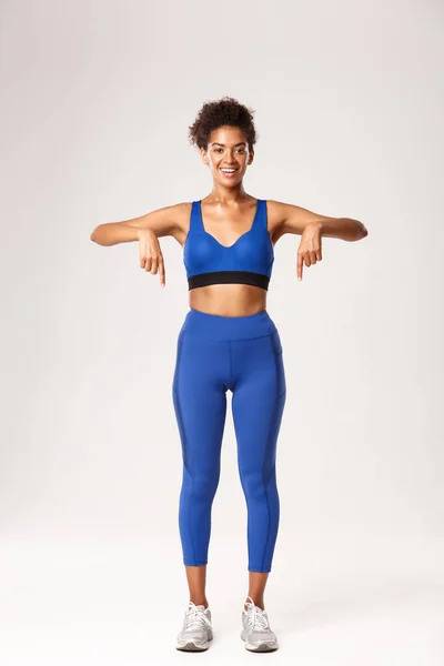 Full length of beautiful african-american fitness girl, wearing sports clothing, pointing fingers down and smiling with happy face, showing advertisement or logo, white background — Foto de Stock