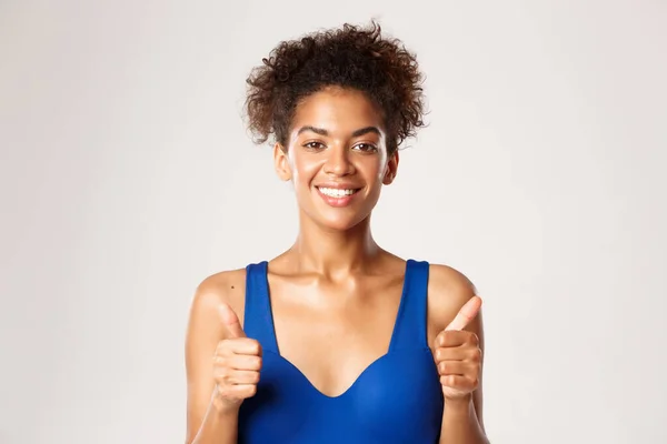 Attraktiv och frisk afrikansk-amerikansk fitness kvinna i blå sport behå, visar tummen upp i godkännande och leende, står över vit bakgrund — Stockfoto