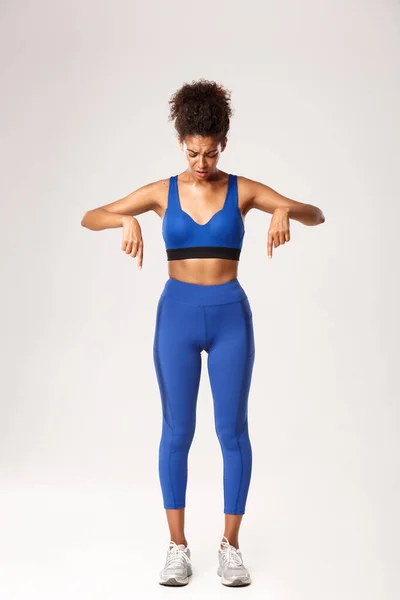 Mavi spor giyim giyen, üzgün, üzgün, üzgün bir ifadeyle, beyaz arka planda duran, parmaklarını aşağı doğru gösteren, üzgün, Afro-Amerikan fitness kızı. — Stok fotoğraf